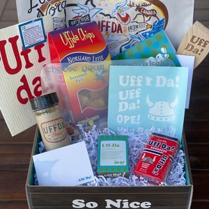 May include: A gift box filled with various items, including a bag with the text "L'Etoile du Nord - That's French for Star of the North!" and a red bag with the text "Uffda Chips - Potato Lefse". The box also contains a small bag of seasoned salt, a jar of seasoning, a small blue and white soap bar, a red tin of bandages, and a white card with the text "Uff Da!" The box is decorated with a wooden sign that says "So Nice".
