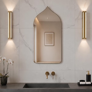 May include: A pointed arch wall mirror with a thin frame, flanked by two vertical gold wall sconces. Below, a dark gray rectangular sink features a gold faucet and handle. A small potted white orchid and a black tray with soap sit nearby.