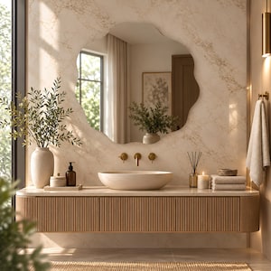 Op de afbeelding: Een badkamermeubel met een aan de muur gemonteerde lichthouten kast met verticale latten, een beige stenen waskom en een gouden kraan. Erboven hangt een grote spiegel met golvende randen die een kamer met een raam weerspiegelt.