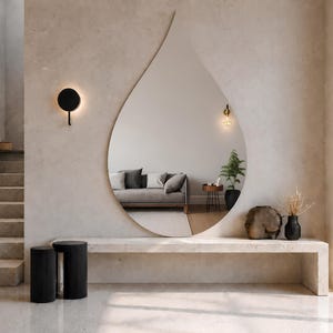Può includere: Un grande specchio da parete senza cornice a forma di goccia d'acqua, montato su una parete beige chiaro sopra una lunga panca rettangolare in pietra. Lo specchio riflette l'interno di un soggiorno con un divano grigio e un tavolino.