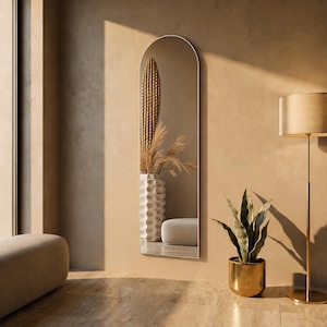 May include: A tall, full-length floor mirror with a thin, arched metal frame. The reflective surface displays a view of a white textured vase containing dried grasses and a tall cactus, alongside a portion of a light-colored, low-profile furniture piece.