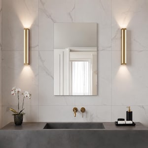 May include: A rectangular wall mirror centered between two vertical, gold-toned wall sconces with integrated LED lighting. The mirror reflects a bright room with white curtains. Below, a dark gray countertop features a sink, gold faucet, and a plant.