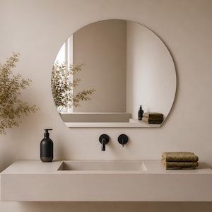May include: A wall-mounted bathroom vanity featuring a beige rectangular sink and a matching countertop. Above the sink is a large, frameless arched mirror. The setup includes a matte black faucet, a black soap dispenser, and folded olive towels.