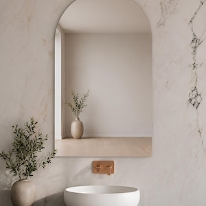 May include: An arched wall mirror with a smooth, frameless edge. The mirror reflects a room with a light-colored wall, a vase with green foliage, and light wood flooring. It is mounted above a white vessel sink and a copper-toned faucet fixture.