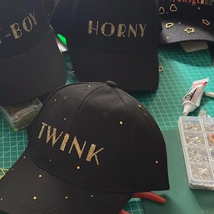 May include: Black baseball caps featuring gold glitter lettering and scattered gold rhinestones. The caps display the words T-BOY, HORNY, and TWINK in a bold font. These headwear items are shown during the crafting process with adhesive and rhinestones.