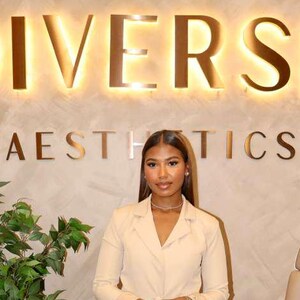 May include: A beige blazer and matching wide-leg trousers worn with a thin metallic necklace. The outfit is displayed in front of a wall featuring the illuminated text DIVERSE AESTHETICS, a potted green plant, and a beige medical treatment chair.