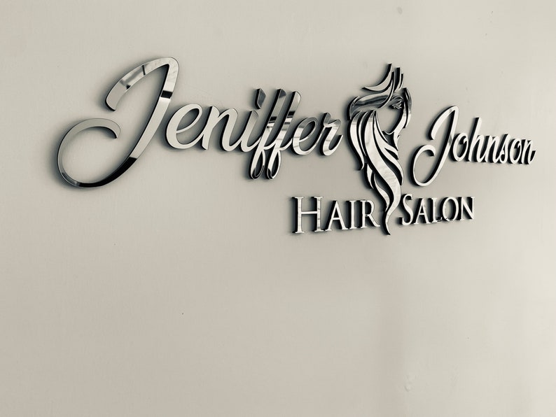 K&ouml;nnte beinhalten: Ein silbernes Schild mit dem Text "Jennifer Johnson Hair Salon" in einer kursivschriftlichen Schriftart. Das Schild zeigt ein stilisiertes Bild von Frauenhaaren.