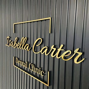 May include: Gold lettering on a black wall reads "Isabella Carter Dental Clinic" with a gold frame around the name.