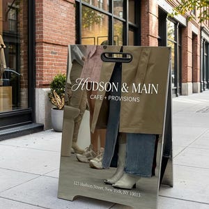 May include: A-frame sign with the text "HUDSON & MAIN CAFE + PROVISIONS" in white. The sign reflects the image of people wearing various outfits. The address "123 Hudson Street, New York, NY 10013" is printed at the bottom.