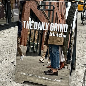 May include: A reflective metal sandwich board sign with the text "THE DAILY GRIND Matcha" in white. The sign reflects a street scene with people and buildings. The address "1234 MAIN ST., SANTA MONICA, CA 90401" is at the bottom.