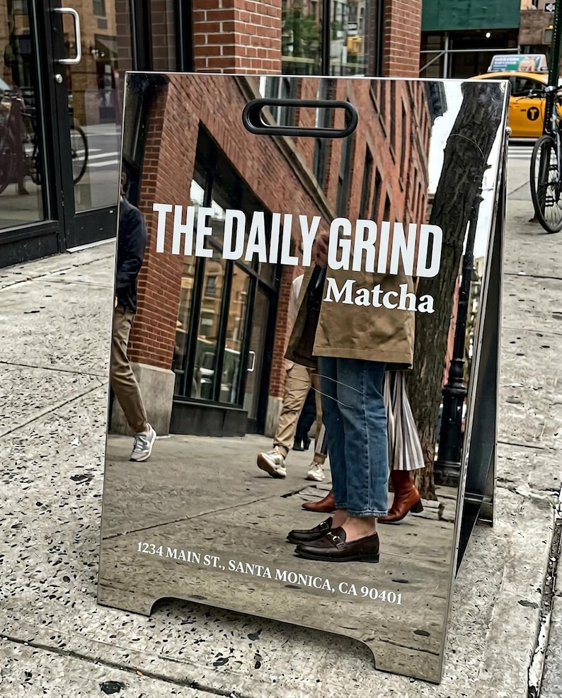 May include: A reflective metal sandwich board sign with the text "THE DAILY GRIND Matcha" in white. The sign reflects a street scene with people and buildings. The address "1234 MAIN ST., SANTA MONICA, CA 90401" is at the bottom.