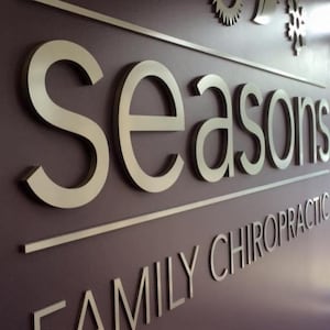 K&ouml;nnte beinhalten: Ein silbernes Metallschild mit dem Wort "Seasons" in erhabenen Buchstaben. Unter dem Wort "Seasons" befindet sich der Text "Family Chiropractic" in kleineren erhabenen Buchstaben.