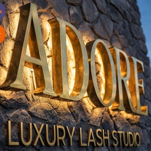 May include: A three-dimensional sign with gold-colored metal lettering reading ADORE LUXURY LASH STUDIO mounted on a textured gray stone wall. The large letters are backlit with warm light, creating a glowing effect against the stone surface.