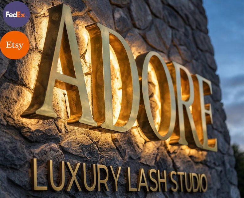 May include: A three-dimensional sign with gold-colored metal lettering reading ADORE LUXURY LASH STUDIO mounted on a textured gray stone wall. The large letters are backlit with warm light, creating a glowing effect against the stone surface.