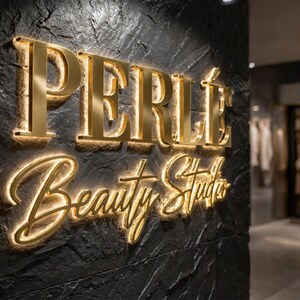 May include: A three-dimensional wall sign featuring the gold-colored text PERL&Eacute; Beauty Studio mounted on a dark, textured, charcoal-colored stone surface. The letters are illuminated from behind, creating a glowing halo effect around each character.