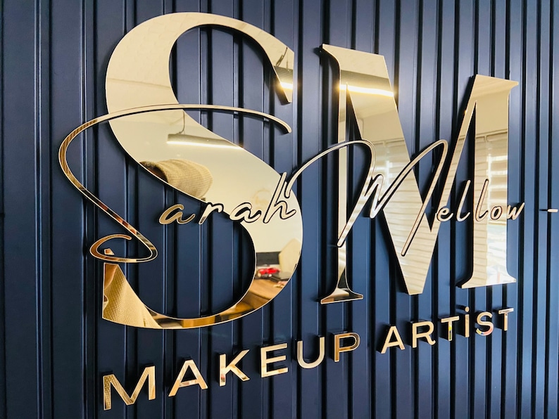 May include: Gold mirrored letters spelling "SM" with the text "Sarah Mellow Makeup Artist" below. The letters are mounted on a dark blue wall.