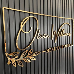 May include: Gold metal sign with the text "Olivia Williams - Beauty Studio" in a cursive font. The sign is mounted on a black wall with vertical grooves.