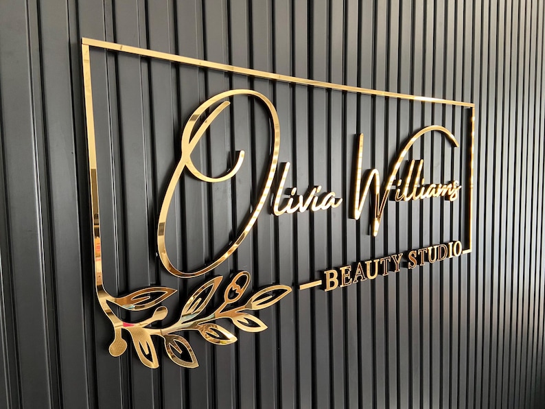 May include: Gold metal sign with the text "Olivia Williams - Beauty Studio" in a cursive font. The sign is mounted on a black wall with vertical grooves.