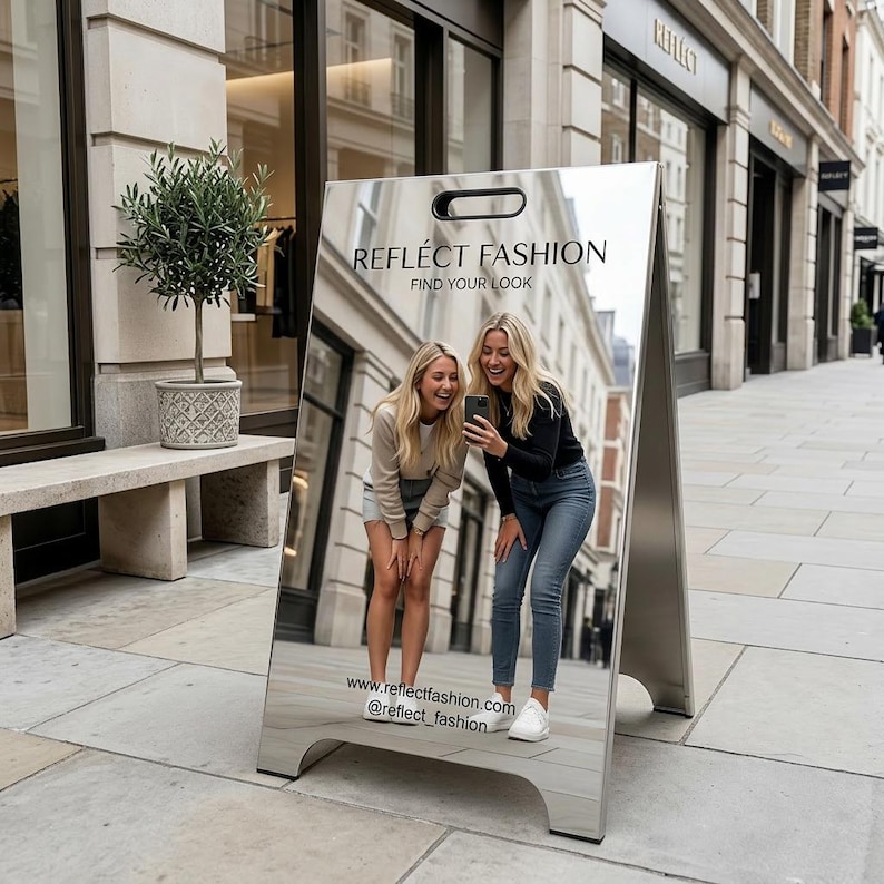 May include: A-frame sign with the text "REFLECT FASHION" and "FIND YOUR LOOK." The sign reflects a street scene with two women looking at a phone. The sign is silver and has a handle at the top. The website and Instagram handle are also displayed.
