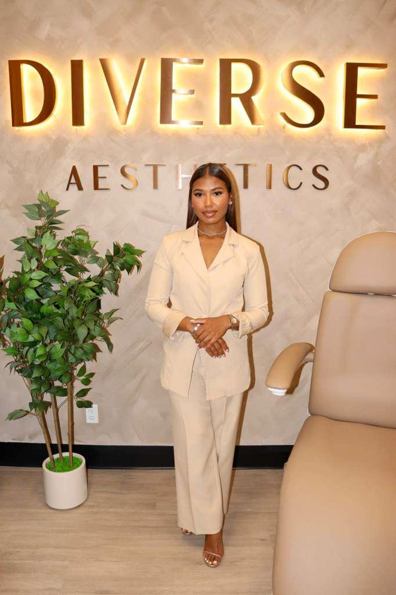 May include: A beige blazer and matching wide-leg trousers worn with a thin metallic necklace. The outfit is displayed in front of a wall featuring the illuminated text DIVERSE AESTHETICS, a potted green plant, and a beige medical treatment chair.