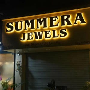 May include: A storefront sign with illuminated gold lettering that reads SUMMERA JEWELS, with the smaller text Fine Diamond Jeweller below. The sign is mounted above a dark, horizontal-slatted metal security shutter at night.