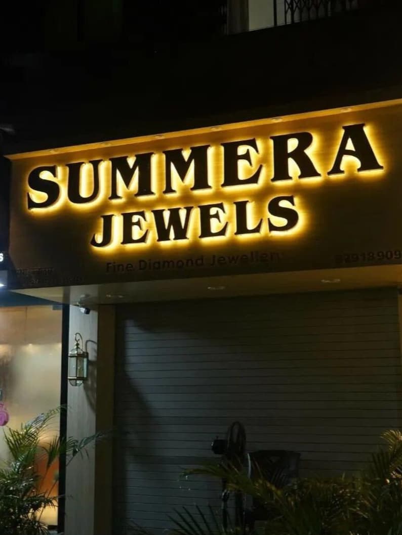 May include: A storefront sign with illuminated gold lettering that reads SUMMERA JEWELS, with the smaller text Fine Diamond Jeweller below. The sign is mounted above a dark, horizontal-slatted metal security shutter at night.