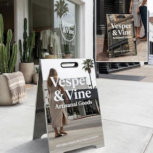 May include: A silver A-frame sign with the text "Vesper & Vine Artisanal Goods" in white. The sign reflects the surroundings, including a person and a storefront. The address "1105 Abbot Kinney Blvd, Venice, CA 90291" is printed at the bottom.