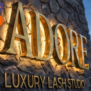 May include: A three-dimensional sign with gold-colored metal lettering reading ADORE LUXURY LASH STUDIO mounted on a textured gray stone wall. The large letters are backlit with warm light, creating a glowing effect against the stone surface.