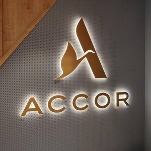 May include: A wall-mounted sign featuring the gold-colored Accor logo with a stylized bird icon integrated into the letter A. The lettering and icon are backlit, creating a glowing halo effect against a gray, perforated metal wall panel.