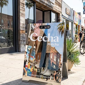 May include: A-frame sign with the text "C&eacute;cha Matcha" in white, reflecting the street scene. The sign displays images of clothing and footwear. The address "1501 Abbot Kinney Blvd, Venice, CA 90291" is printed at the bottom.