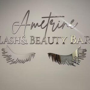 K&ouml;nnte beinhalten: Ein silbernes Schild mit dem Text "Ametrine Lash & Beauty Bar" in einer kursiven Schrift. Zwei silberne Wimpern befinden sich auf beiden Seiten des Textes.