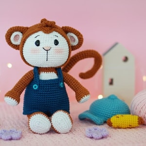 Może przedstawiać: Szydełkowa lalka małpki z brązowymi i białymi detalami, w niebieskich ogrodniczkach. Na zdjęciu znajduje się niebieska czapka, żółty banan i kulka różowej przędzy. Tło jest w kolorze jasnoróżowym. Widoczne są również inne szydełkowe przedmioty.