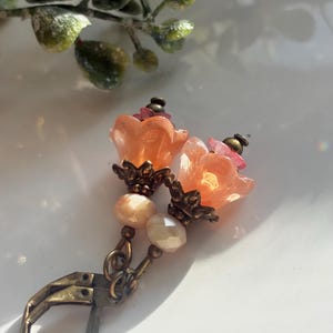 May include: A pair of dangle earrings featuring translucent peach-colored flower-shaped beads, small faceted cream-colored beads, and dark bronze-toned metal findings, including ornate bead caps and secure lever-back closures for attachment.