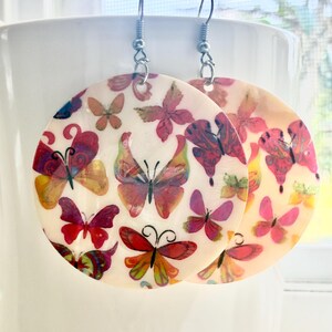 May include: A pair of round, flat earrings featuring a colorful butterfly pattern in shades of pink, red, orange, and yellow on a cream-colored background. The earrings are suspended from silver-toned metal hooks and have a smooth, glossy finish.