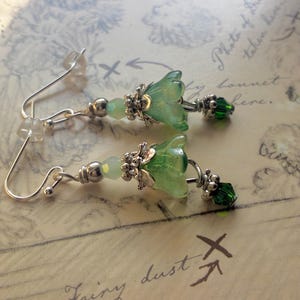 May include: A pair of dangle earrings featuring silver-toned hooks, small pale green beads, and silver-toned metal flower caps. Each earring includes a translucent light green bell-shaped glass flower and a small, faceted dark green crystal bead.