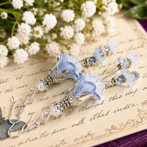 May include: A pair of dangle earrings featuring blue and white bell-shaped flower beads, silver-toned metal filigree caps, small white pearls, and clear crystal accents. The earrings are attached to silver-toned metal ear wires for pierced ears.