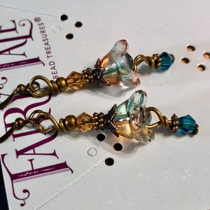 May include: A pair of dangle earrings featuring a central flower-shaped glass bead with iridescent blue and gold tones. The earrings are accented with smaller amber and teal crystal beads, antique brass-toned wire, and metal findings.