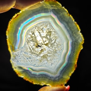 May include: A slice of agate with a rainbow iridescent sheen. The agate is a light brown color with a white center and a dark brown edge.