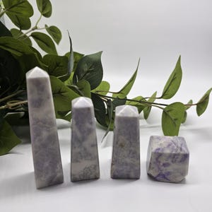 May include: Four polished lepidolite crystal towers of varying heights, each featuring a smooth, faceted surface with a marbled pattern of soft lavender, light purple, and white hues, arranged in a row from tallest to shortest on a surface.