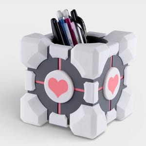 Puede incluir: Un organizador de escritorio blanco, gris y rosa con forma de Cubo de Compañía del videojuego Portal. El cubo tiene dos corazones rosas en los lados y contiene varios bolígrafos y lápices.