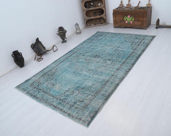 May include: A rectangular, distressed-look rug in shades of teal and grey. The rug features a central teal field with a faded, patterned design and a dark grey border. The rug is placed on a white floor.