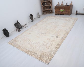 Può includere: Un tappeto rettangolare beige con un intricato motivo floreale. Il tappeto ha un bordo dettagliato ed è posato su un pavimento bianco. Oggetti decorativi e mobili sono visibili sullo sfondo.