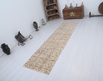 Puede incluir: Una alfombra larga y rectangular de color beige con un estampado floral. La alfombra está colocada sobre un suelo de madera de color claro. Varios objetos decorativos antiguos están en el fondo, incluyendo un mueble de madera y una escultura de metal.
