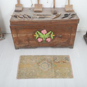 May include: A small, rectangular, vintage rug with a faded, geometric pattern in shades of beige, yellow, and blue. The rug is placed on a white floor in front of a wooden chest with a floral design.