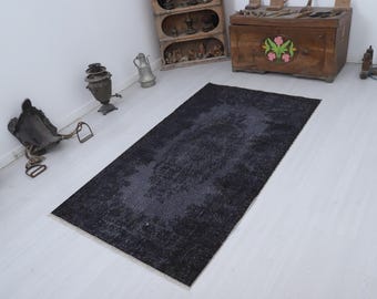 Puede incluir: Una alfombra rectangular gris oscuro con un patrón descolorido y desgastado. La alfombra está colocada sobre un suelo de madera de color claro. El diseño de la alfombra presenta un área central de color más claro, creando un contraste sutil. La alfombra es un revestimiento decorativo para el suelo.