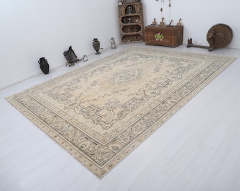 May include: A large, rectangular beige rug with an intricate, dark brown floral pattern. The rug has a detailed border and is placed on a light-colored floor. The rug is the focal point of the room, with antique furniture and decor in the background.