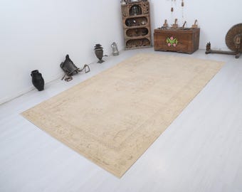 May include: A rectangular, beige-colored rug with an intricate, faded pattern. The rug is placed on a light-colored wooden floor. The rug's design features a central field with a detailed border. The rug is a large size, suitable for a living room or bedroom.