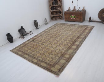 May include: A rectangular beige rug with a repeating circular pattern in shades of brown and red. The rug has a detailed border and is placed on a white floor. Decorative items are in the background.