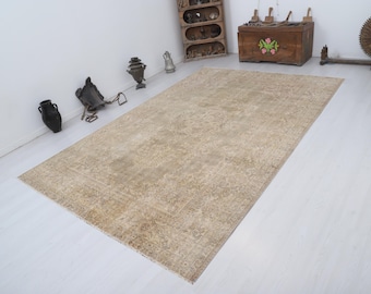 May include: A rectangular, beige-toned rug with an intricate, faded floral pattern. The rug is placed on a white floor, and various antique decorative items are visible in the background, suggesting a vintage or bohemian aesthetic.
