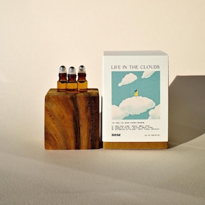 May include: A perfume set with the text "LIFE IN THE CLOUDS" on the box. The box features an illustration of a person on a cloud. Three small amber glass bottles sit on a wooden block. The set is labeled as an Eau de Parfum.